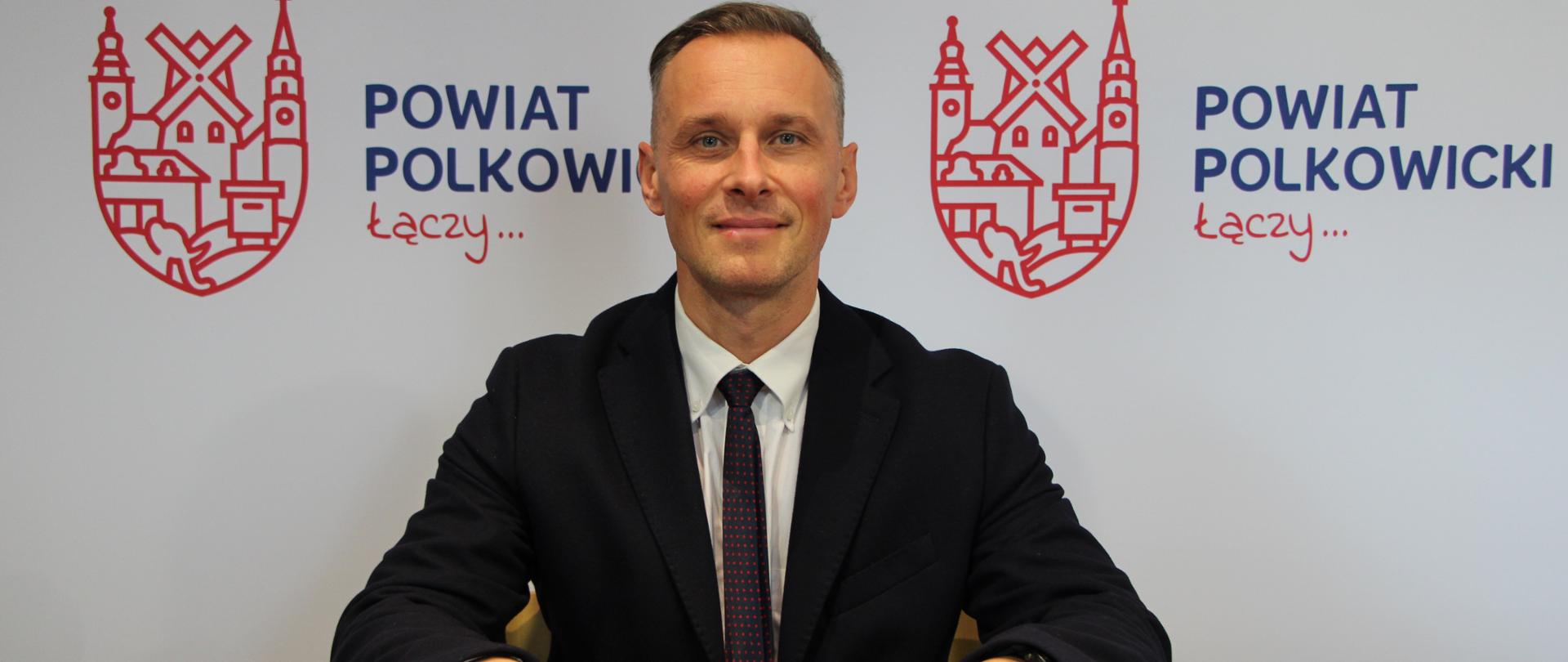 Uśmiechnięty Mężczyzna w garniturze i krawacie siedzi przy stole, przed nim leży notes.
