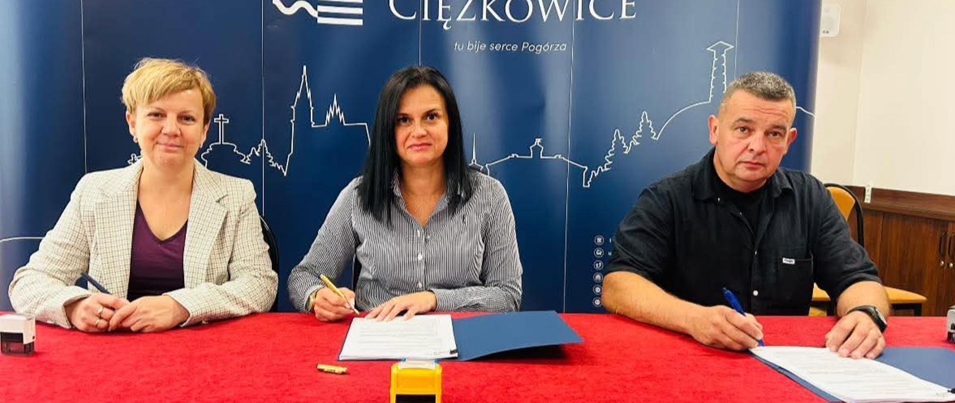Trzy osoby siedzące na tle granatowej ściany z napisem gmina Ciężkowice