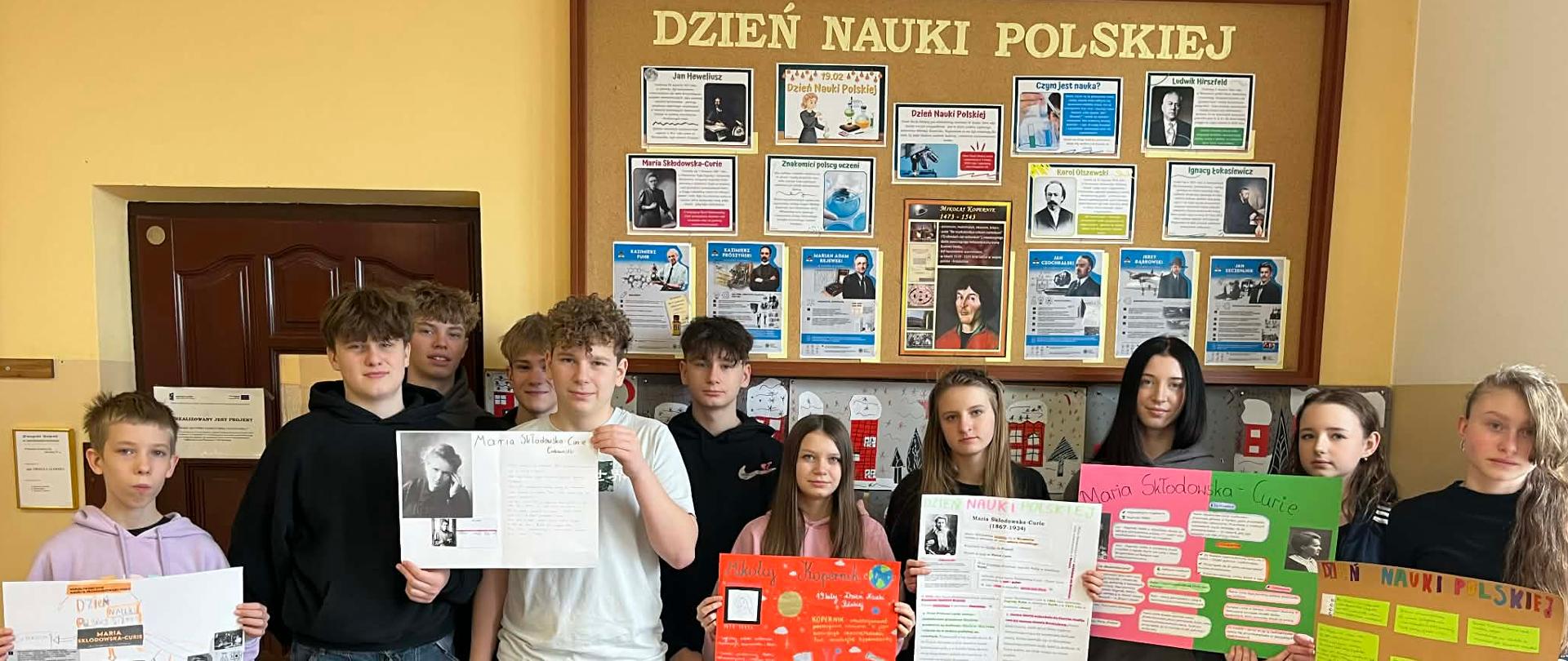 Młodzież klasy 8 na tle tablicy szkolnej poświęconej obchodom Dnia Nauki Polskiej prezentuje swoje plakaty
