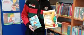 Uczeń trzyma w ręku książkę i dyplom, za nim regały biblioteczne