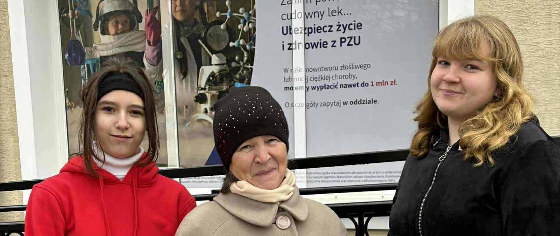 Dwie uczennice klasy ósmej pozują do wspólnego zdjęcia ze starszą Panią. Kobieta trzyma w dłoni opaskę odblaskową.