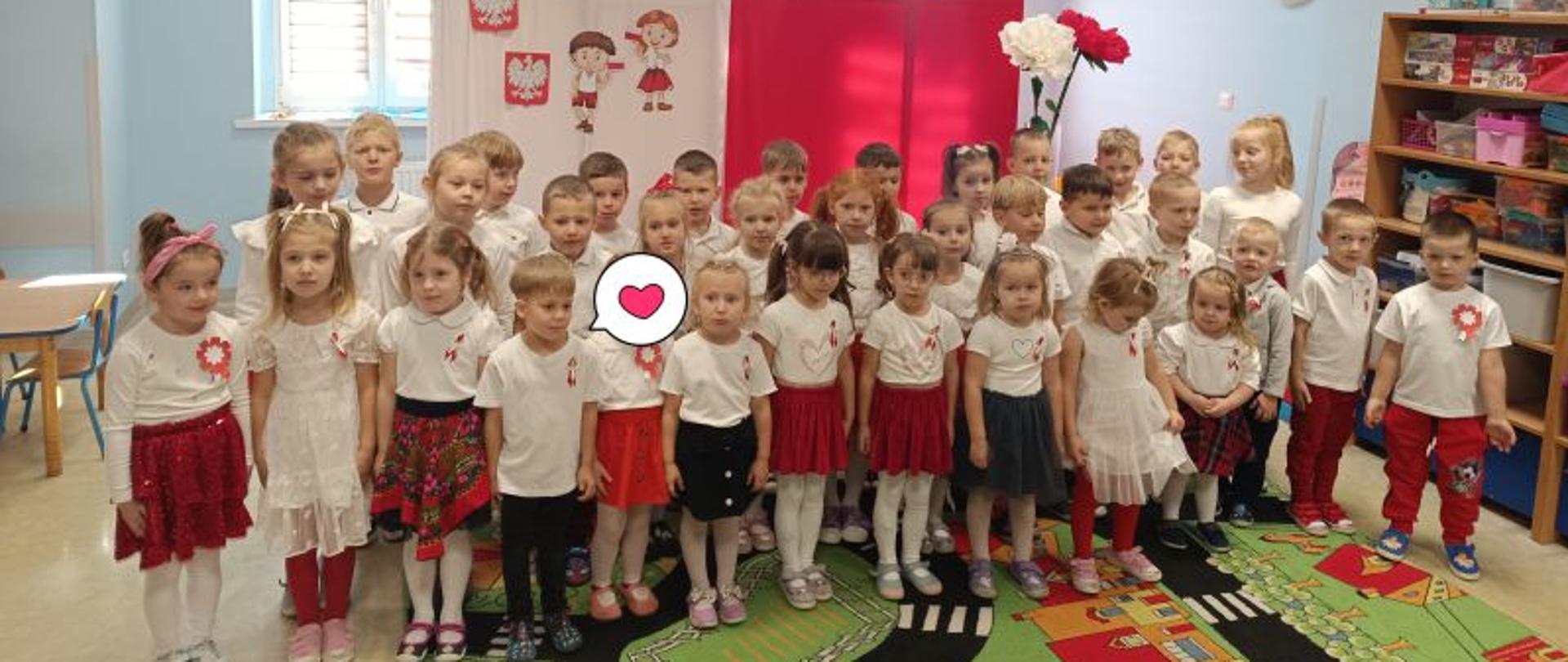 Dzieci w strojach biało-czerwonych stoją na tle ścianki w kolorach flagi Polski, z boku dwa duże kwiaty w kolorach białym i czerwonym. 