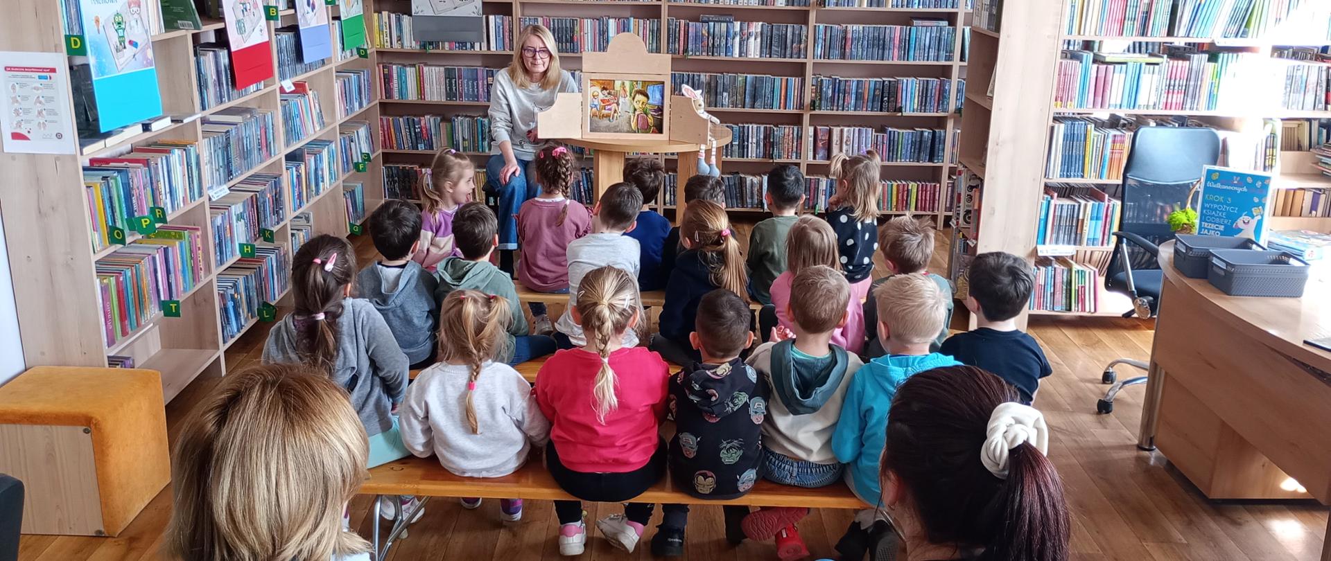 Zdjęcie przedstawia dzieci siedzące na ławeczkach i słuchające opowiadanej przez bibliotekarkę bajki w formie teatrzyku kamishibai