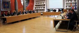 Duża sala konferencyjna po dwóch stronach stoły a przy nich siedzący mężczyźni i kobiety. W centralnej części stół przy którym również siedzą trzy osoby. Na ścianie centralnej herby sześciu gmin powiatu polkowickiego oraz herb powiatu. 