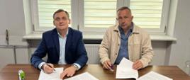Od lewej Wójt Gminy Wietrzychowice oraz przedstawiciel firmy, którzy siedzą przy stole i podpisują dokumenty. W tle okno przysłonięte jasną żaluzją