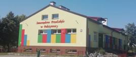 Zdjęcie przedstawia budynek z napisem „Samorządowe Przedszkole w Pokrzywnicy”. Fasada budynku jest w kolorze jasnokremowym, ozdobiona pionowymi pasami w kolorach czerwonym, zielonym, niebieskim i żółtym. W górnej części znajdują się dwa małe okna, a poniżej trzy większe. Przed wejściem znajduje się utwardzony plac, a po prawej stronie stoją zaparkowane rowery. Budynek jest ogrodzony, a w tle widoczne są drzewa.
