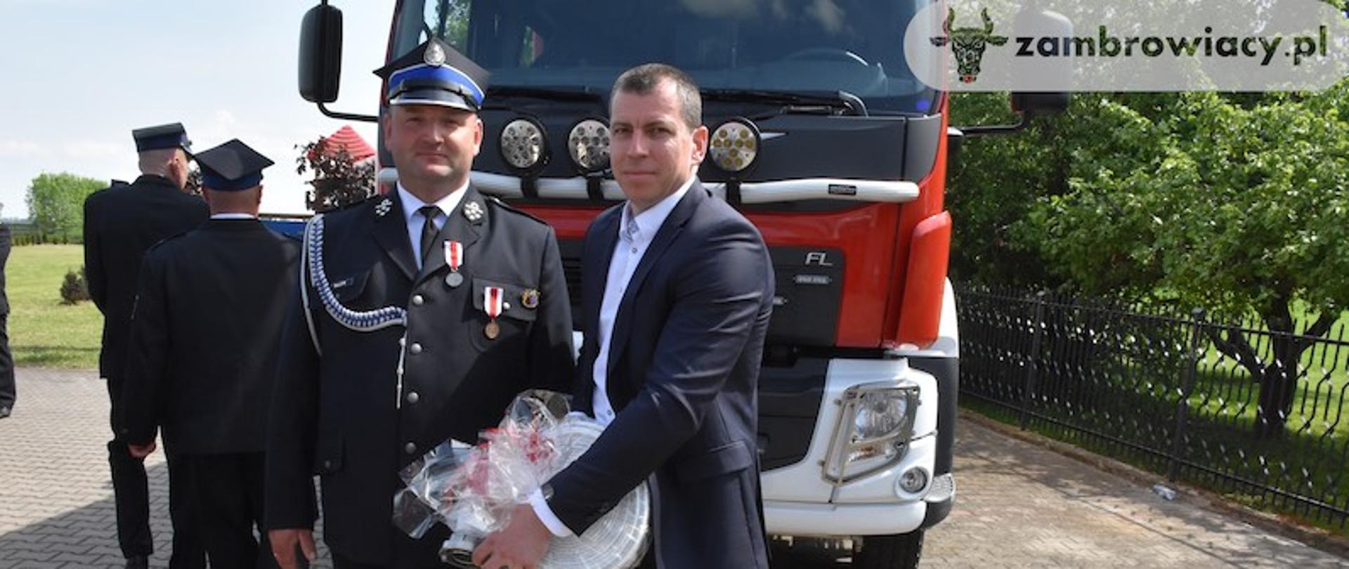 Radny Powiatu Zambrowskiego Sławomir Worosilaka przekazuje na ręce prezesa OSP Pechratka Polska strażacki wąż, w tle znajduje się czerwony wóz strażacki