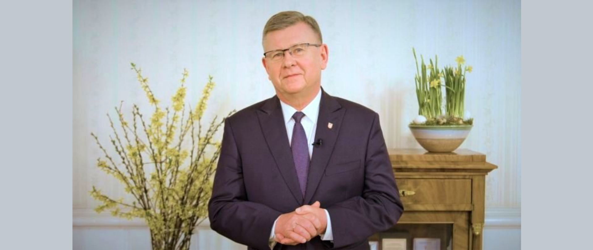 Mężczyzna na środku zdjęcia za nim donica z kwitnącymi narcyzami oraz bukiet kwitnącej forsycji.
