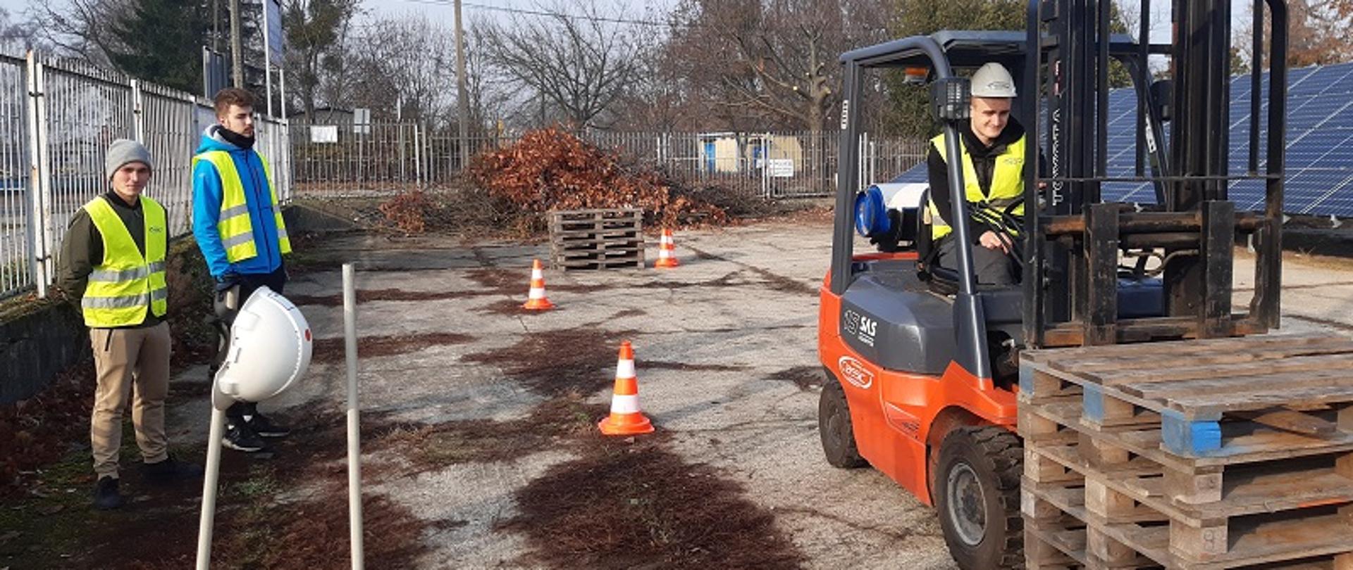 Zdjęcie przedstawia ucznia klasy logistycznej Zespołu Szkół Powiatowych w Przasnyszu podczas manewrowania wózkiem widłowym. Po lewej dwóch nauczycieli bacznie się przygląda oraz poddaje ucznia ocenie. 