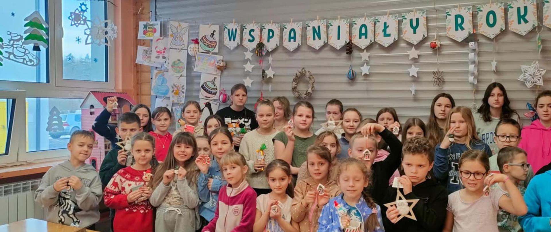 Na tle dekoracji z napisem "Wspaniały rok" stoją uczniowie biorący udział w konkursie, trzymają w rękach przygotowane przez siebie ozdoby choinkowe