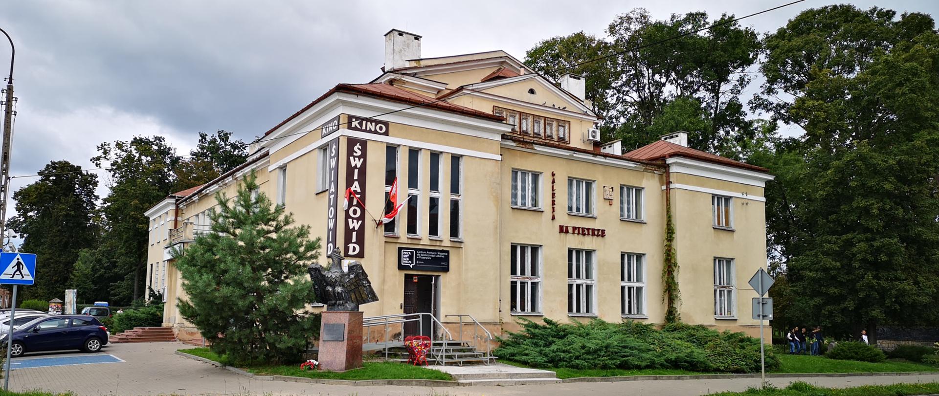Zdjęcie przedstawia Miejski Dom Kultury w Przasnyszu. Po lewej stronie parking, po prawej Park Miejski imienia Tadeusza Kościuszki. Widok od ulicy Świętego Stanisława Kostki.