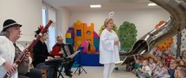 Zdjęcie prezentuje dzieci biorące udział w audycji muzycznej oraz teatrzyku. Przed grupą siedzących dzieci występują aktorzy