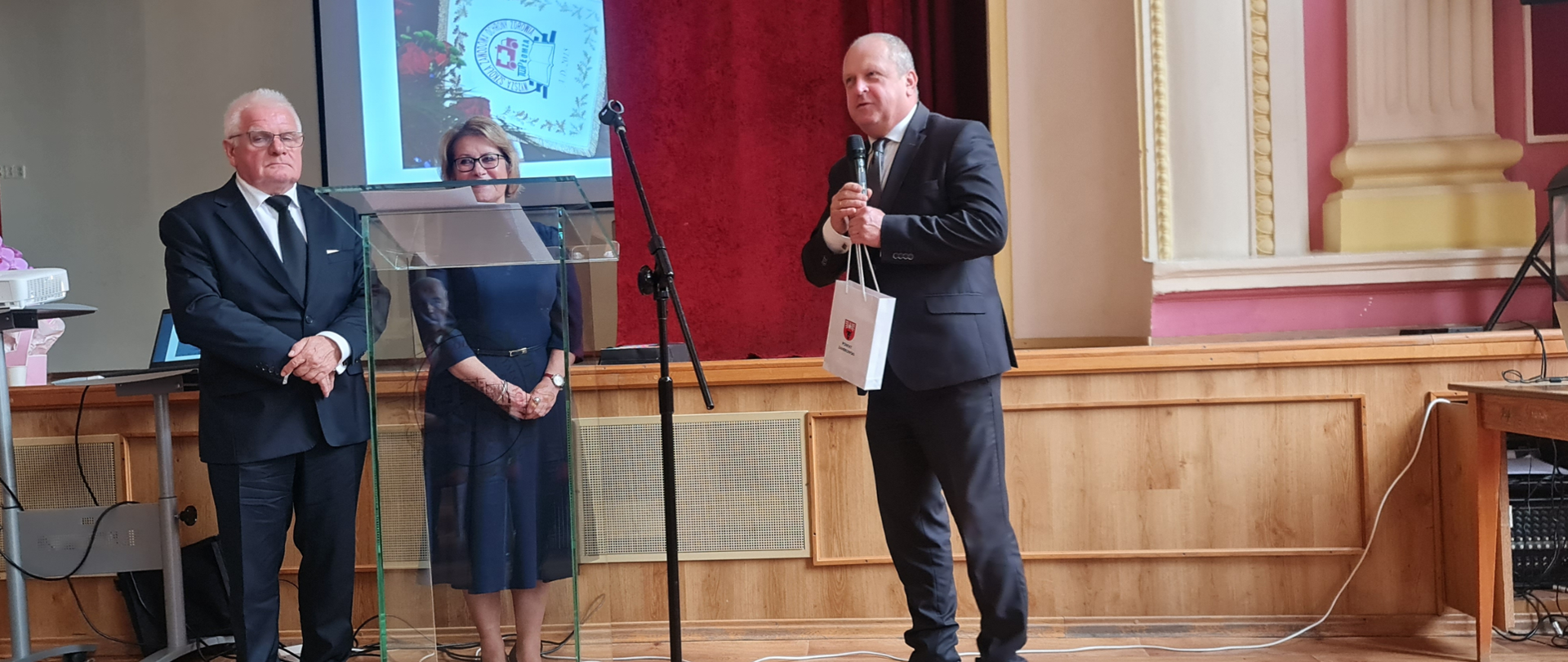 Na zdjęciu widać wicestarostę zambrowskiego przemawiającego do publiczności. Występuje on podczas uroczystości 25-lecia Oddziału Regionalnego Towarzystwa Wiedzy Powszechnej (TWP) w Łomży, która odbyła się 11 października 2025 roku. Wydarzenie miało miejsce w auli II LO w Łomży. Na zdjęciu widoczne są również inne osoby, a w tle widać ekran projekcyjny.