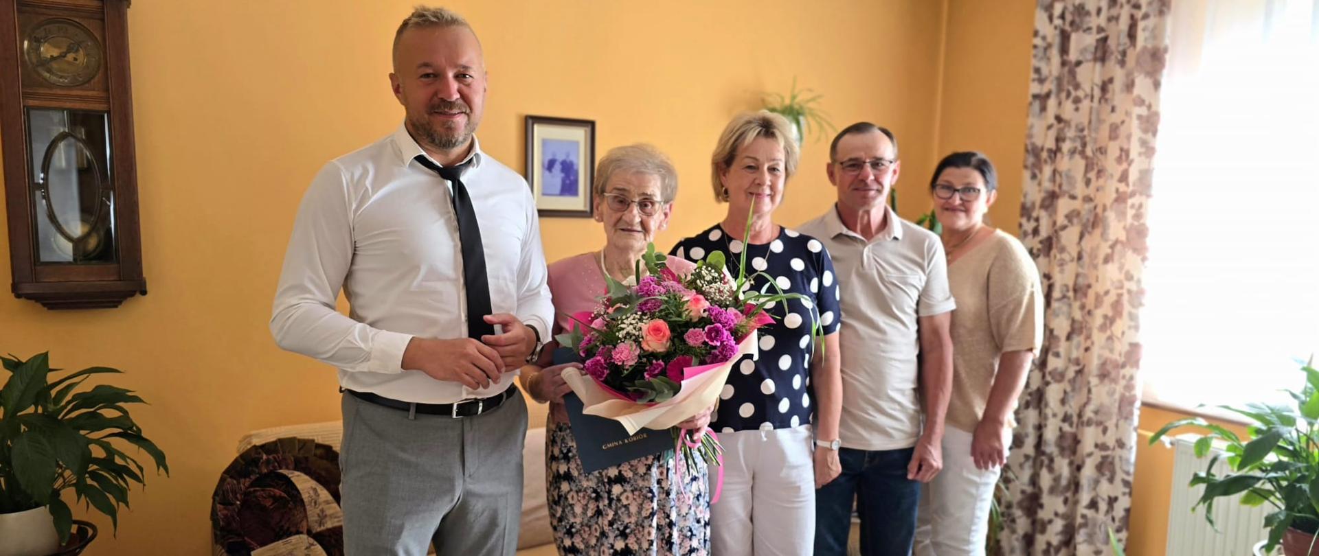 Grupa osób, w tym jubilatka z bukietem kwiatów, pozują do zdjęcia