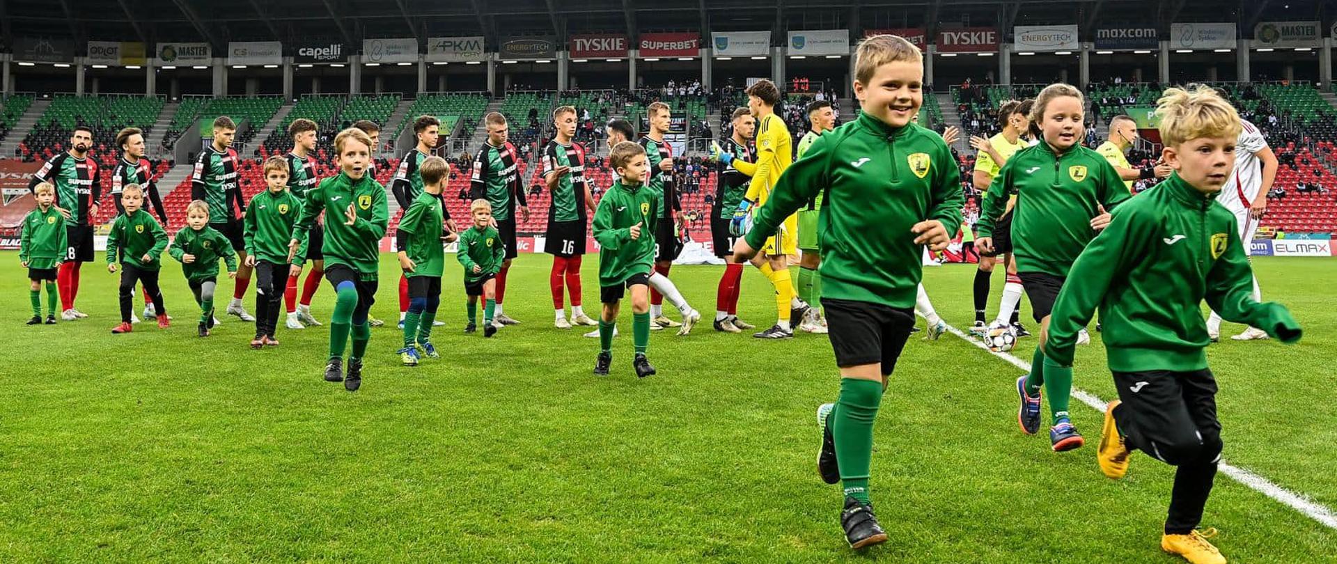 Dzieci w zielonych stroją biegną na murawie boiska. Za nimi stoją piłkarze.