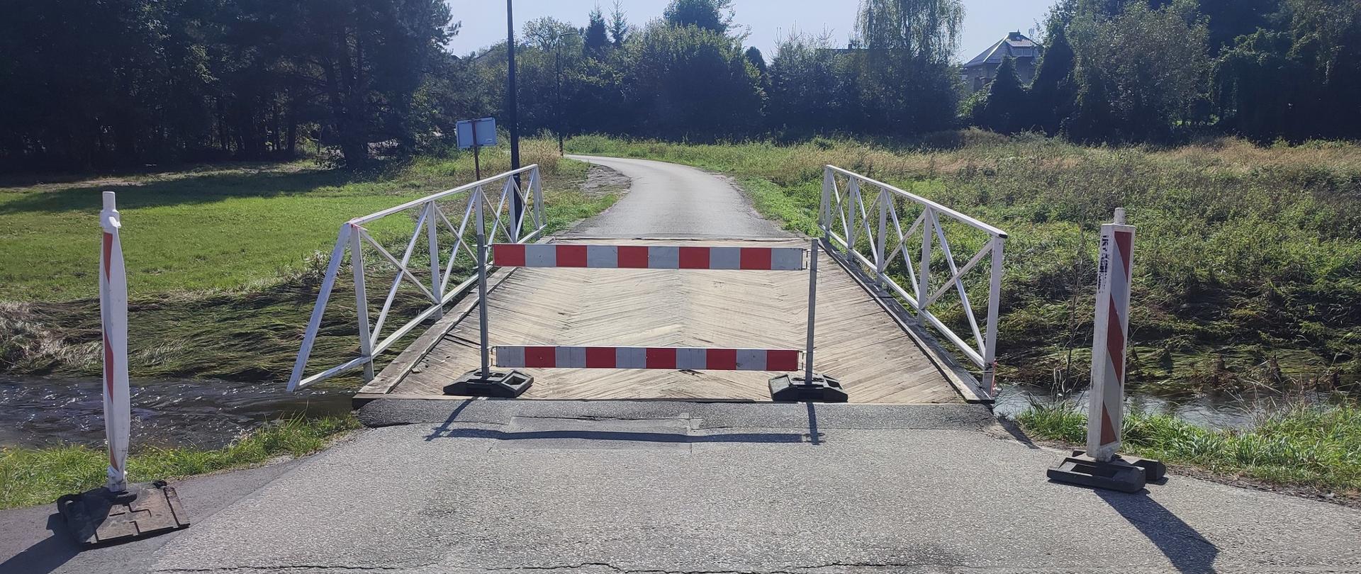 Drewniany most nad małą rzeką z metalowymi białymi barierkami. Przed mostem stoi biało czerwona bariera uniemożliwiająca wjazd. 