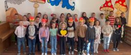 Uczniowie z klas I-III stoją w trzech szeregach i składają babciom i dziadkom życzenia, kilkoro z nich ma żółte opaski z czerwonym serduszkiem na głowie, inni trzymają w dłoniach czerwone serduszka 