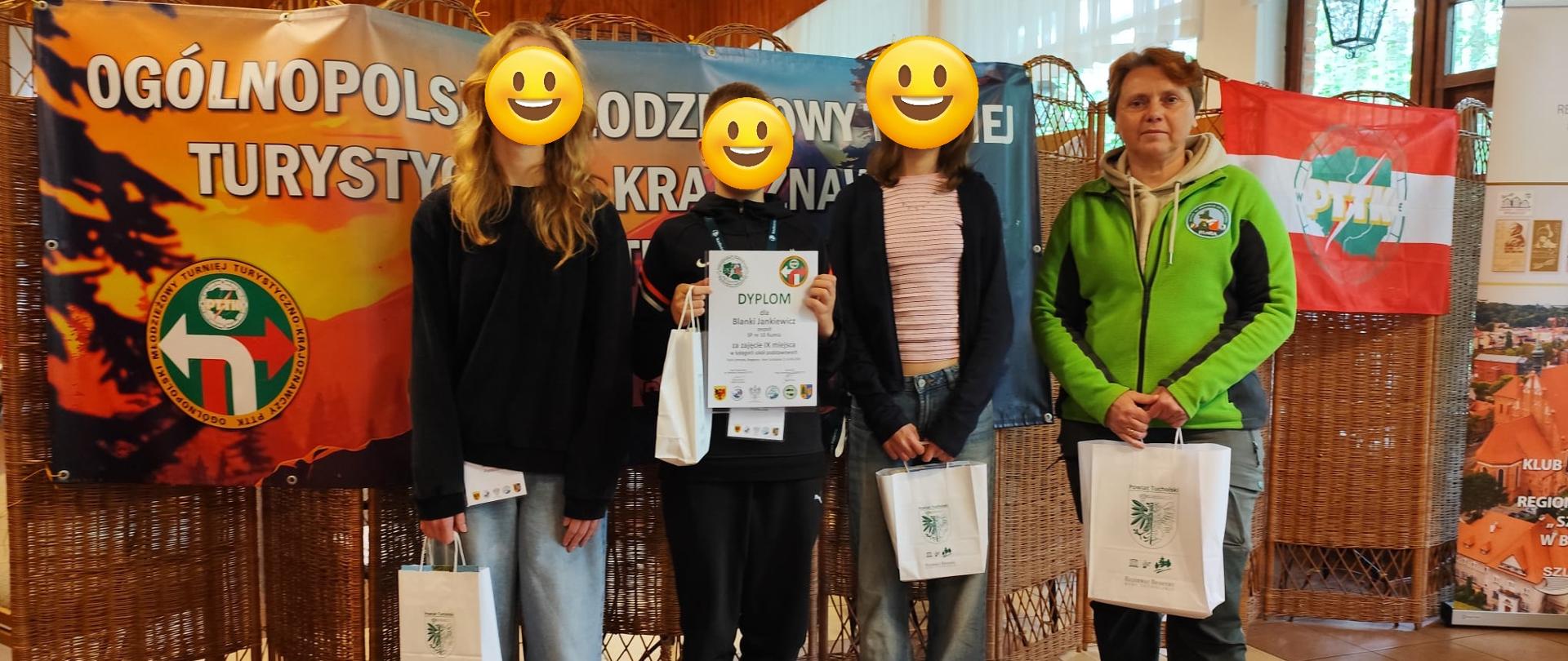 Na górze zdjęcia sufit w kolorze brązowym z lampami. Z prawej strony okna z białymi zasłonami. Na środku zdjęcia sylwetki czterech osób ustawionych obok siebie, w dłoniach trzymające dyplomy oraz papierowe torebki w kolorze białym. Za osobami baner w kolorach pomarańczowo niebieskich z białymi literami. Podłoga w kolorze jasnobrązowym.