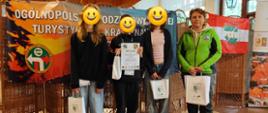 Na górze zdjęcia sufit w kolorze brązowym z lampami. Z prawej strony okna z białymi zasłonami. Na środku zdjęcia sylwetki czterech osób ustawionych obok siebie, w dłoniach trzymające dyplomy oraz papierowe torebki w kolorze białym. Za osobami baner w kolorach pomarańczowo niebieskich z białymi literami. Podłoga w kolorze jasnobrązowym.