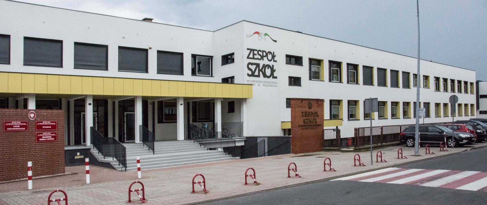 Biały budynek szkoły Zespołu Szkół im. Narodów Zjednoczonej Europy w Polkowicach. Na budynku logo szkoły. Przed budynkiem tablica informacyjna. Na początku zdjęcia widoczna ulica i przejście dla pieszych.