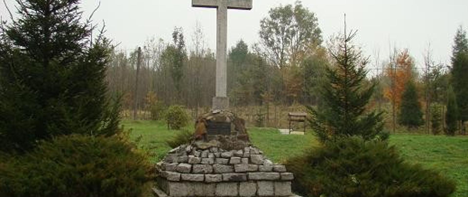 widoczny pomnik upamiętniający wydarzenie krzyż schodki po lewej i prawej widoczne nasadzenia w tle las młodnik widoczna studnia