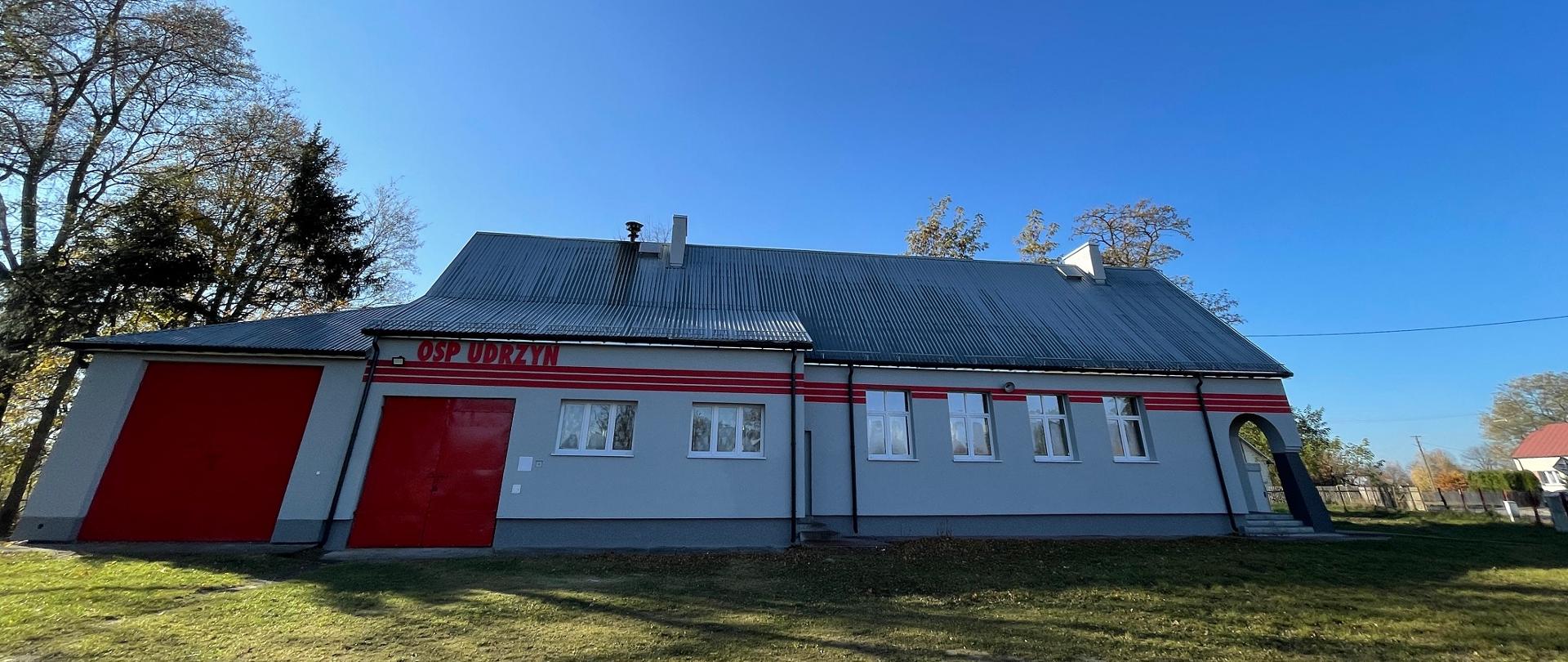 Remiza OSP Udrzyn z nową elewacją w jasnym kolorze. Na elewacji namalowano trzy czerwone poziome pasu oraz napis OSP Udrzyn. Od frontu znajdują się 2 bramy wjazdowe oraz 6 okien.