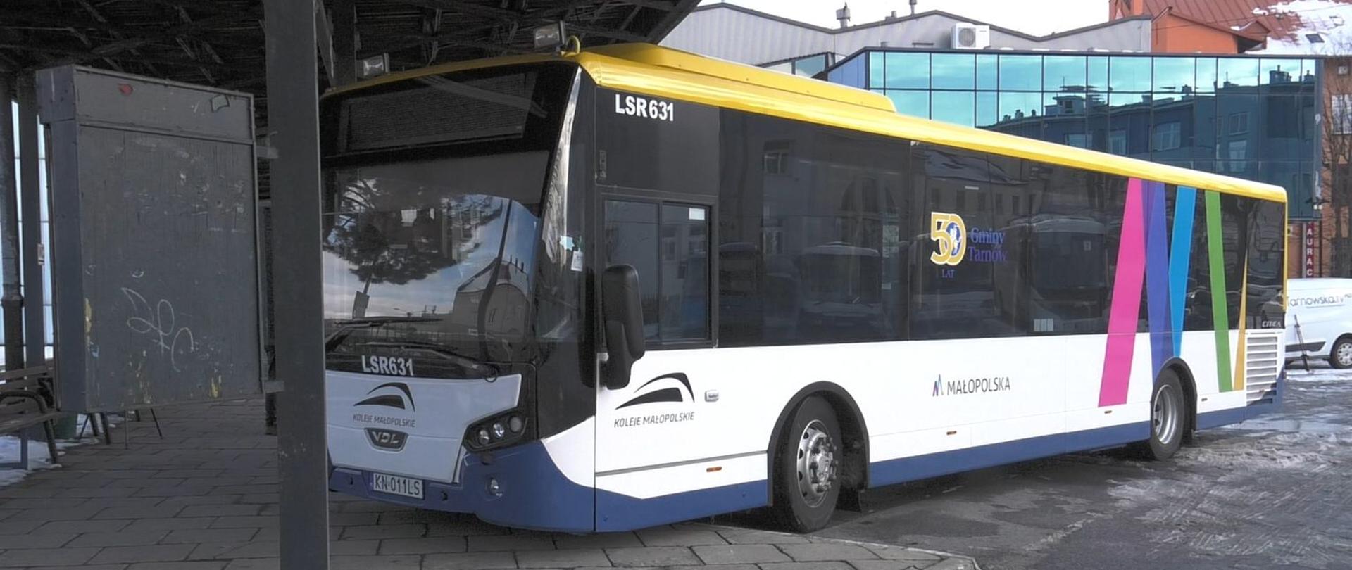 Autobus Kolei Małopolskich linii dowozowej, biało-żółty z kolorowymi pasami, zaparkowany na przystanku przy dworcu w miejskiej zabudowie.