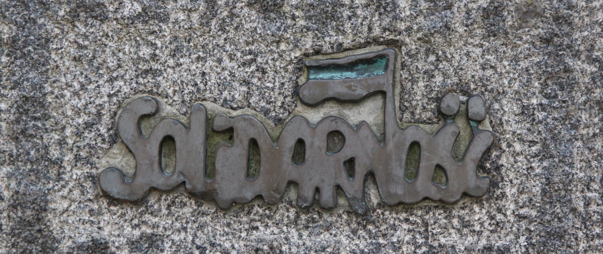 Na fotografii widzimy fragment kamiennego słupa na którym umieszczono metalowy przestrzenny napis "Solidarność"