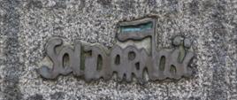 Na fotografii widzimy fragment kamiennego słupa na którym umieszczono metalowy przestrzenny napis "Solidarność"