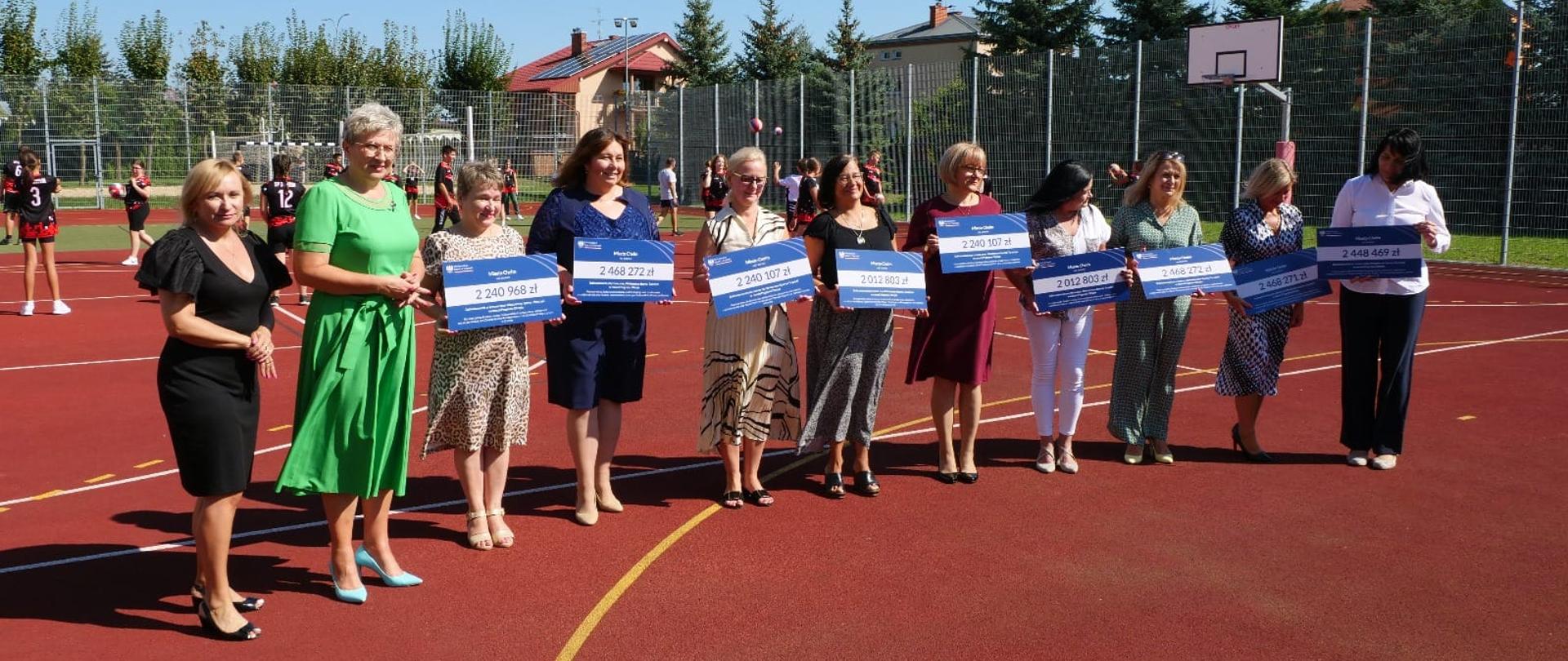 Zdjęcie przedstawia 11 osób stojących w rzędzie na boisku szkolnych trzymających w rękach tabliczki. 