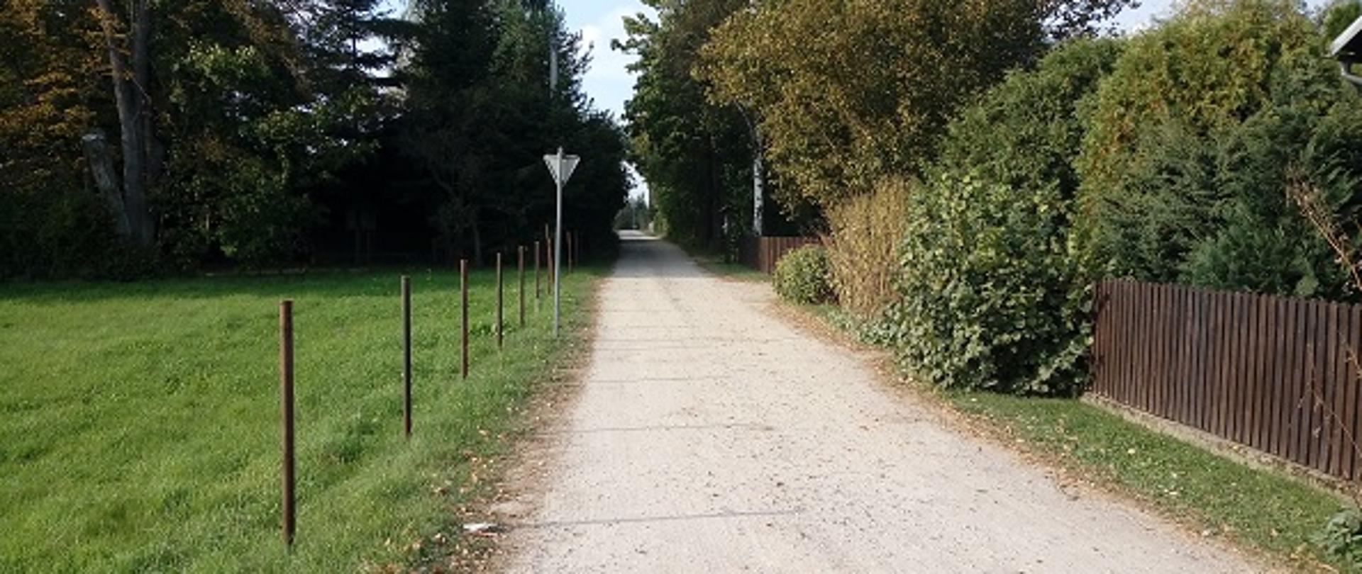 Na zdjęciu widoczna droga gruntowa w miejscowości Poręba Kocęby. Po lewej stronie drogi znajduje się łąka. Po prawej stronie widać zabudowania oraz płoty ogradzające posesję.