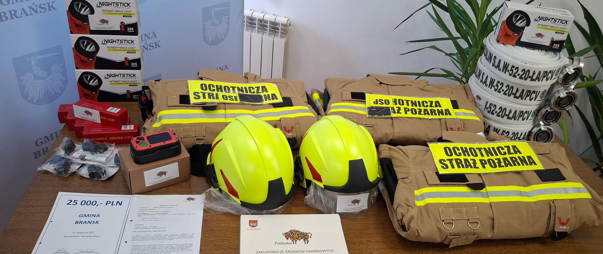 Na drewnianym stole ustawiono nowy sprzęt strażacki: trzy żółte hełmy ochronne, kilka kompletów beżowego umundurowania z odblaskami i naszywkami „Ochotnicza Straż Pożarna”, przewody pożarnicze, latarki oraz inne akcesoria techniczne. Obok leży dokument z kwotą „25 000 PLN” oraz tabliczka informująca o zakupie ze środków finansowych województwa. W tle widać stojak z granatową ścianką oznaczoną napisem „Gmina Brańsk” i herbami.