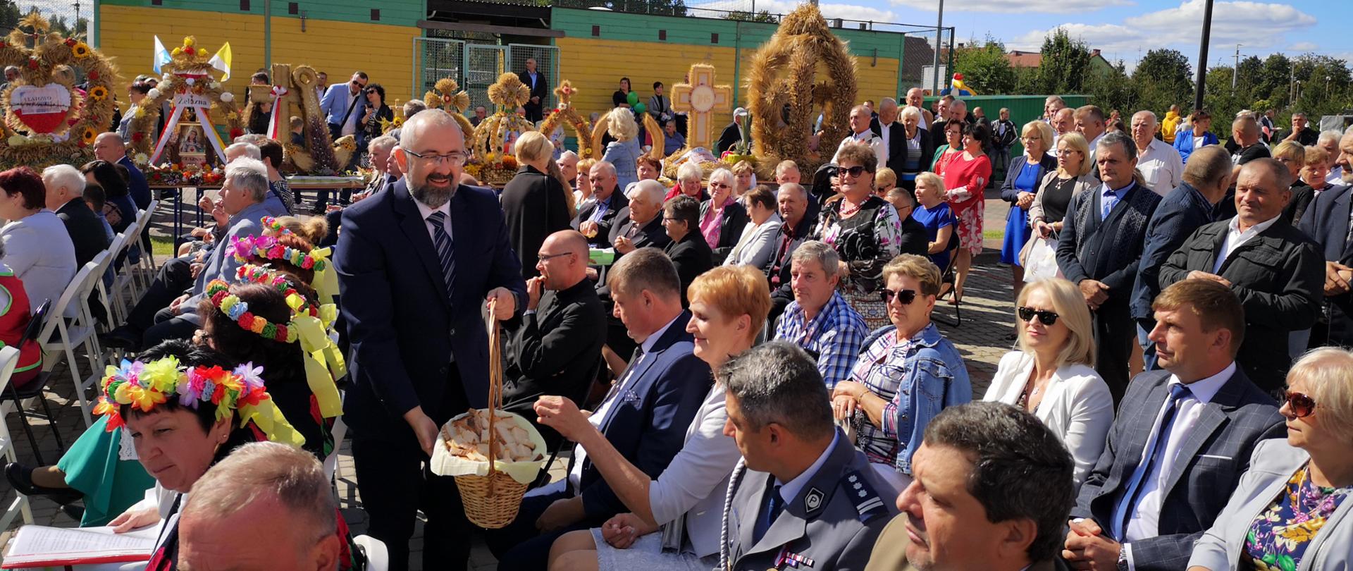 Starosta Przasnyski Krzysztof Bieńkowski dzieli się chlebem dożynkowym z gośćmi oraz uczestnikami Dożynek Powiatowo Gminnych w Jednorożcu. Na fotografii Starosta Przasnyski wyciąga koszyk z chlebem do zgromadzonych gości. Dookoła tłum pozostałych uczestników dożynek, w tle stoły na których prezentują się w linii wieńce dożynkowe.