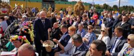 Starosta Przasnyski Krzysztof Bieńkowski dzieli się chlebem dożynkowym z gośćmi oraz uczestnikami Dożynek Powiatowo Gminnych w Jednorożcu. Na fotografii Starosta Przasnyski wyciąga koszyk z chlebem do zgromadzonych gości. Dookoła tłum pozostałych uczestników dożynek, w tle stoły na których prezentują się ustawione linii wieńce dożynkowe.