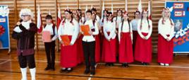Uczniowie klasy 6 ubrani na galowo występują w akademii, po lewej stronie uczeń w stroju króla, z koroną na głowie, z tyłu dziewczynki w białych bluzkach, czerwonych długich spódnicach, w biało-czerwonych wiankach na głowie