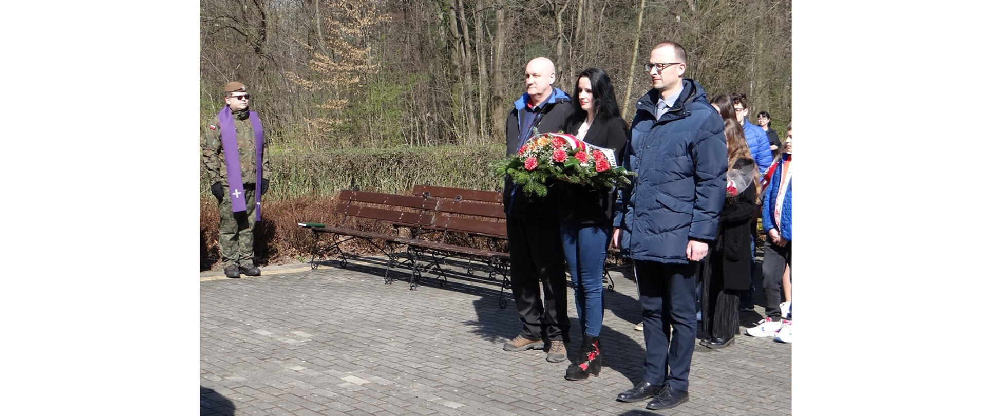 Delegacja z Gminy Hażlach przed pomnikiem ofiar niemieckiego terroru „Pod Wałką” w Cieszynie. W środku trzyosobowej grupy kobieta trzymająca wiązankę kwiatów z szarfą w barwach biało-czerwonych 