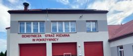 Budynek straży pożarnej z trzema dużymi czerwonymi bramami garażowymi oraz mniejszym czerwonymi drzwiami pośrodku. Nad drzwiami znajduje się napis w języku polskim: „OCHOTNICZA STRAŻ POŻARNA W POKRZYWNICY”. Fasada budynku jest jasnoszara z ciemniejszymi szarymi akcentami wokół okien i drzwi.
