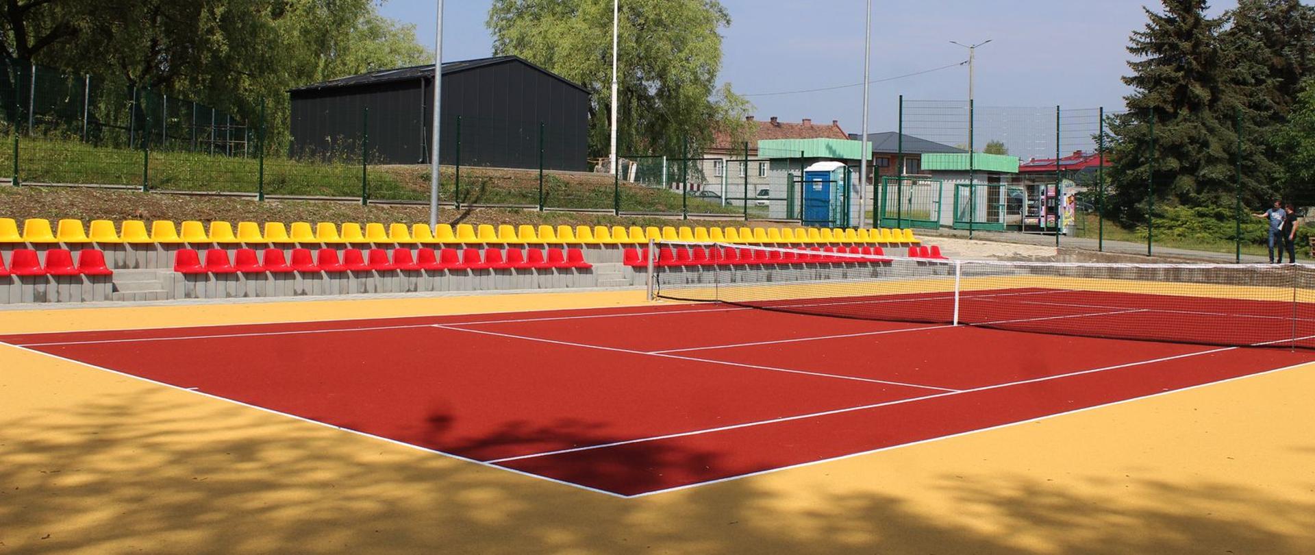 Na pierwszym planie kort tenisowy, w tle krzesełka zółto-czerwone.