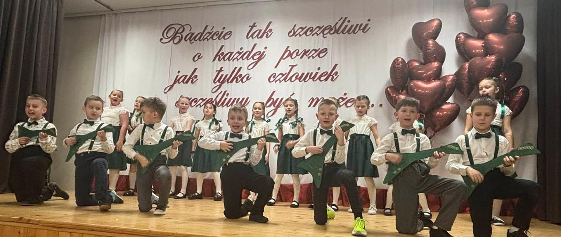 Grupa chłopców w czarnych spodniach, zielonych muchach i szelkach oraz białych koszulach klęczy na jednym kolanie grając na zielonych tekturowych gitarach. Za nimi stoją dziewczynki w zielonych spódniczkach i kokardach we włosach oraz białych bluzeczkach śpiewając. 