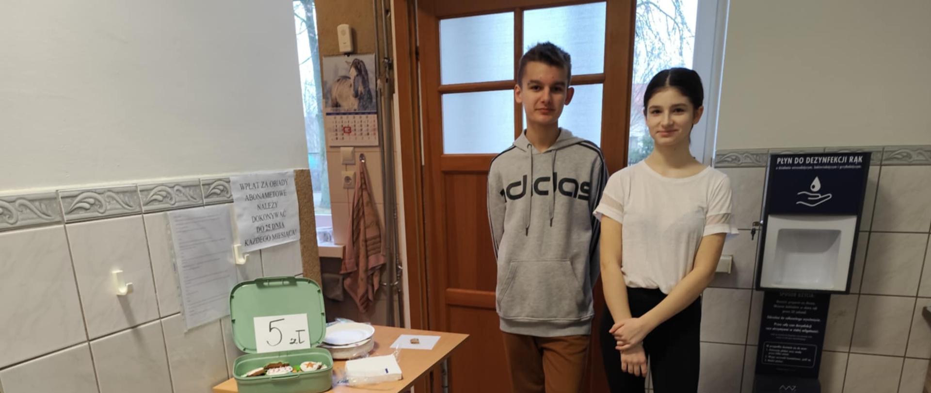 Dwoje uczniów z Samorządu Uczniowskiego - chłopak i dziewczyna pozują do zdjęcia. Przed nimi stoją dwa stoliki z pudełkami i miseczkami pełnymi pierniczków.