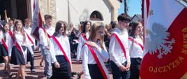 Pochód uczniów w galowych strojach wychodzi z kościoła na zewnątrz. Na czele idą sztandarowi niosący czerwono-biały sztandar szkolny z białym orłem i napisem „Ojczyzna Nauka". Słoneczna pogoda, w tle wierni opuszczający kościół.