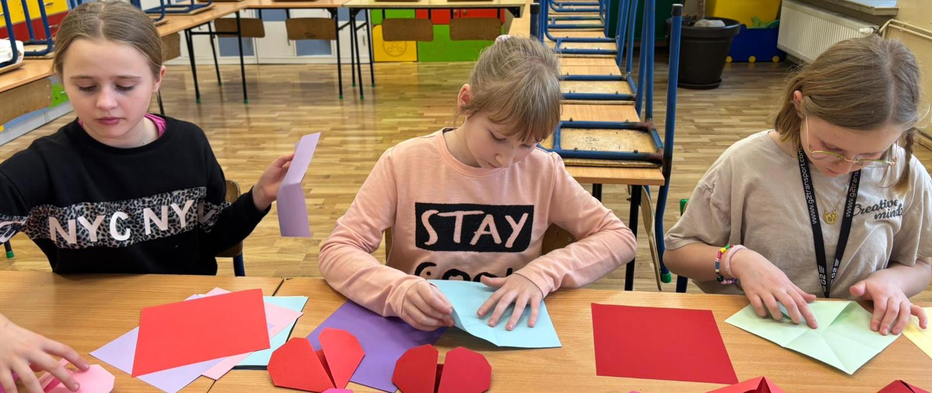 trzy dziewczynki składające z papieru kolorowe serduszka origami