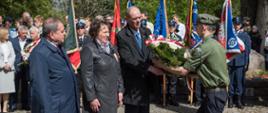 Na fotografii trzyosobowa delegacja Powiatu Sokołowskiego przekazująca wiązankę z biało - czerwonych róż umundurowanemu harcerzowi, który za chwilę złoży ją pod pomnikiem Czynu Niepodległościowego, którego centralną częścią jest okrągły głaz z napisem Bóg, Honor, Ojczyzna.