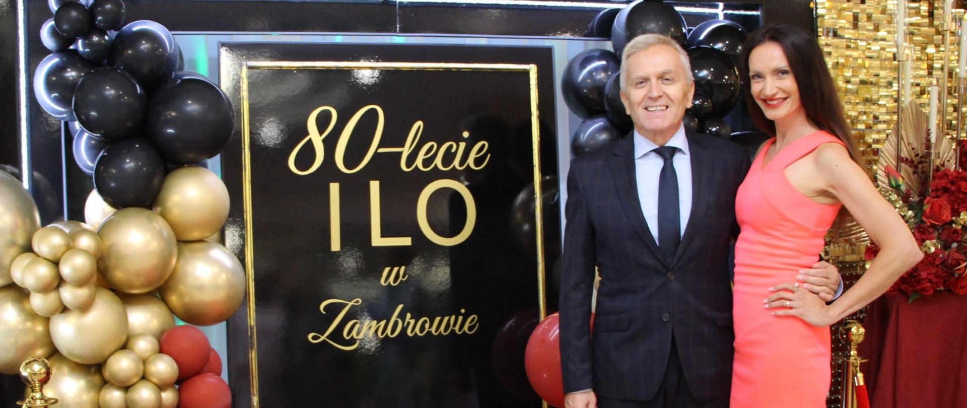 Kobieta i mężczyzna na czerwonym dywanie przed pamiątkową ścianką jubileuszową z napisem „80-lecie I LO w Zambrowie” oraz „Zjazd Absolwentów”. Mężczyzna w ciemnym garniturze i krawacie stoi obok uśmiechniętej kobiety w dopasowanej, koralowej sukience bez rękawów.