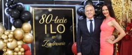 Kobieta i mężczyzna na czerwonym dywanie przed pamiątkową ścianką jubileuszową z napisem „80-lecie I LO w Zambrowie” oraz „Zjazd Absolwentów”. Mężczyzna w ciemnym garniturze i krawacie stoi obok uśmiechniętej kobiety w dopasowanej, koralowej sukience bez rękawów.