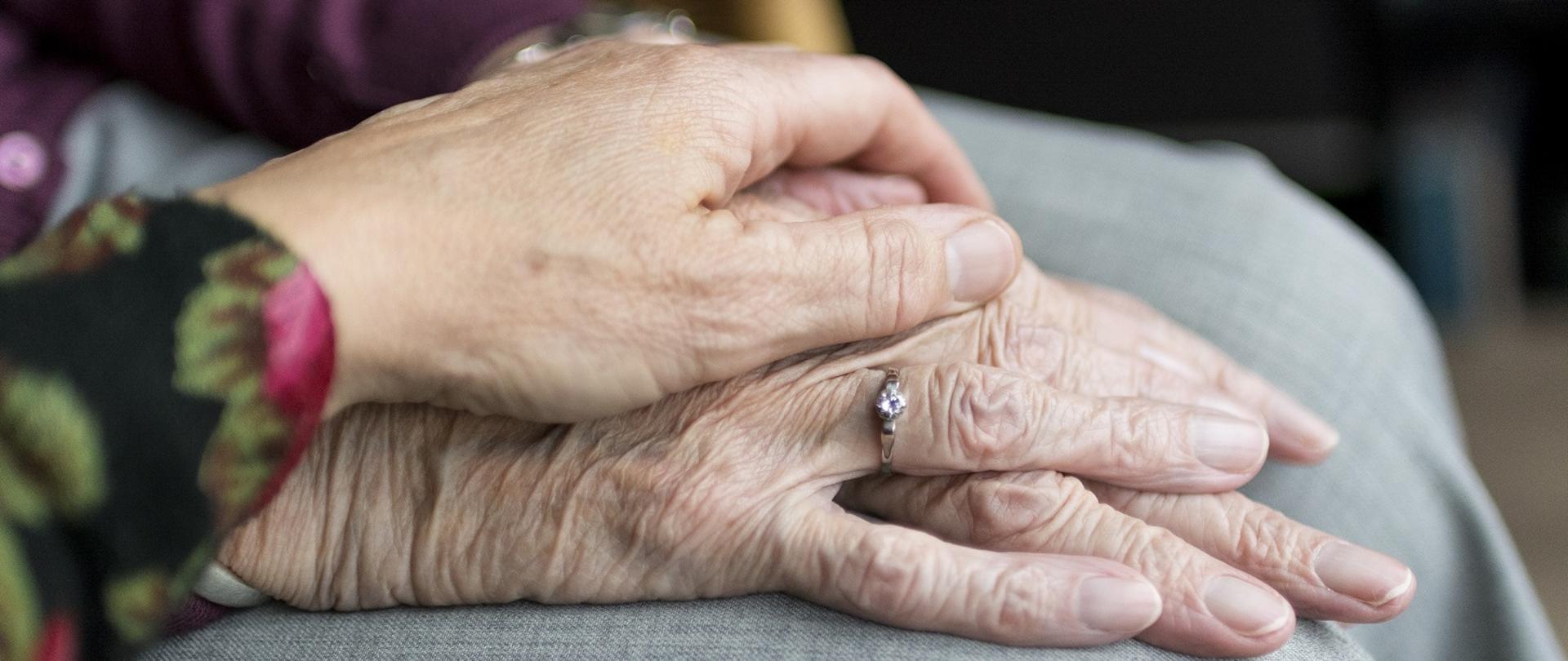 na zdjęciu w kadrze widać złożone na kolanach dłonie starszej osoby, na których z troską położona jest dłoń innej osoby wyrażająca gest wsparcia