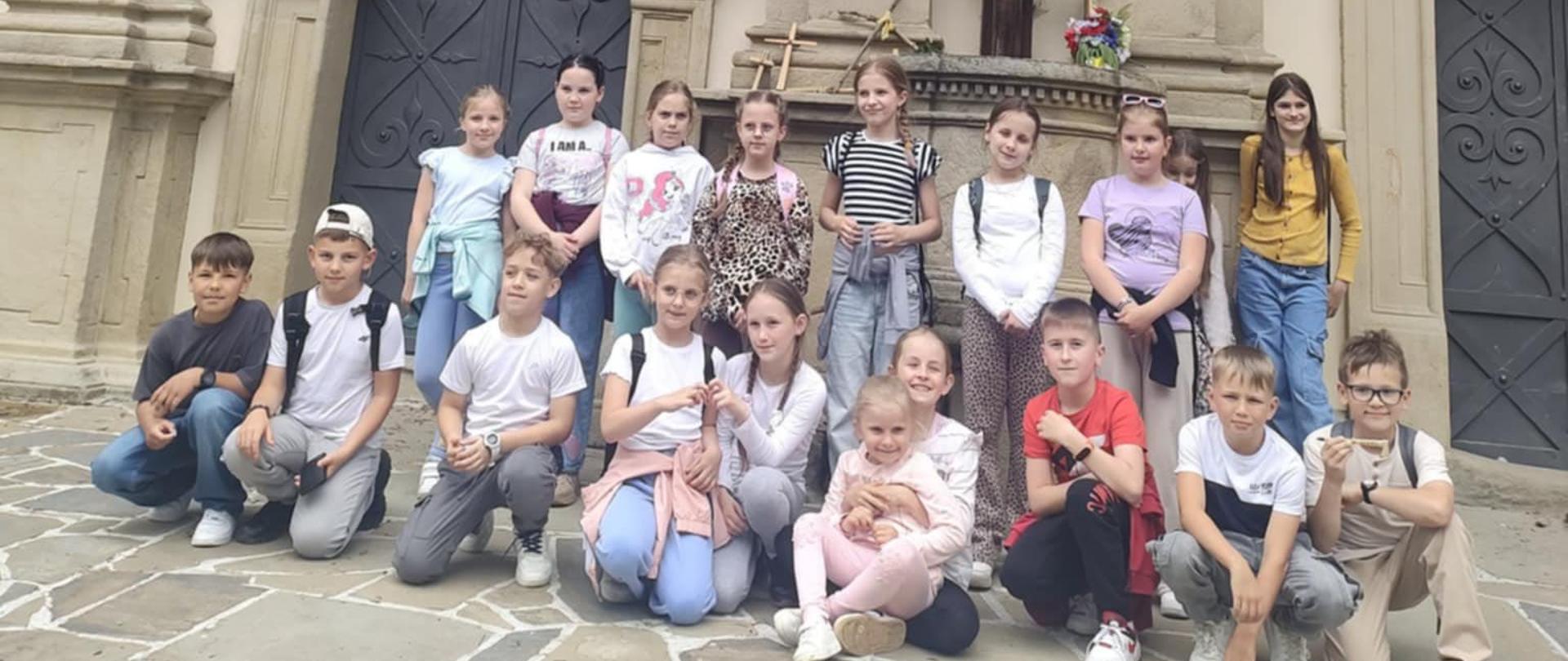 Dzieci stoją przed wejściem do kościoła ozdobionym półokrągłym łukiem, nad nimi krzyż z figurą Jezusa
