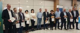 Zdjęcie grupowe hodowców bydła mlecznego z wicestarostą Ryszardem Domańskim. Uczestnicy na fotografii trzymają w dłoniach torebki oraz koperty z logo Powiatu Sokołowskiego. Zdjęcie wykonane na sali bankietowej. W tle rollupy promocyjne firm związanych z hodowlą bydła mlecznego.