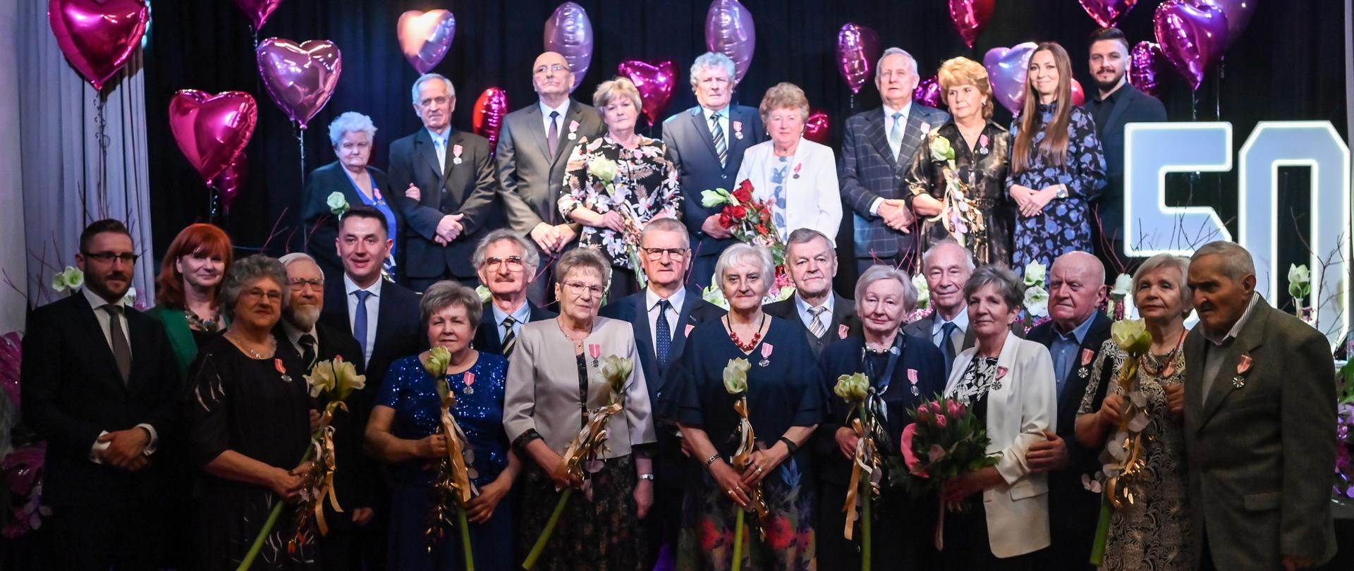 Grupowe zdjęcie par małżeńskich uhonorowanych podczas uroczystości Złotych Godów. Jubilaci stoją na scenie z kwiatami i odznaczeniami, w tle dekoracje i balony w kształcie serc.