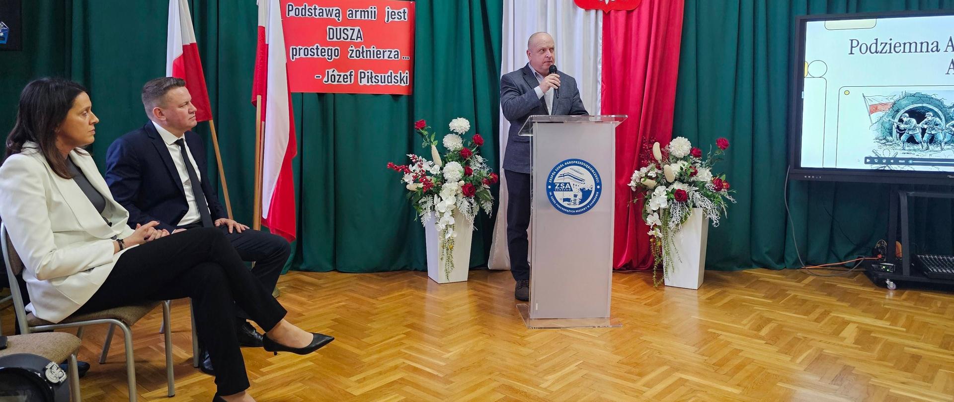 Mężczyzna w szarym garniturze przemawia do mikrofonu, stojąc za przezroczystą mównicą z logo szkoły (ZSA). W tle znajduje się dekoracja w barwach narodowych: zielone zasłony, polska flaga, kontur mapy Polski oraz czerwona tablica z cytatem Józefa Piłsudskiego: „Podstawą armii jest DUSZA prostego żołnierza...”. Po lewej stronie siedzą zaproszeni goście, w tym kobieta w białym żakiecie i mężczyzna w garniturze. Po prawej widać ekran z prezentacją o tytule „Podziemna Armia”.
